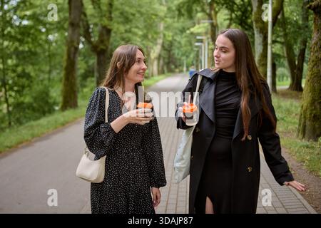 Two young women share lively conversation while holding takeaway coffee cups in tree-lined urban park. Natural daylight and casual attire highlight re Stock Photo