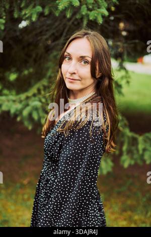 Young woman with straight brown hair and hazel eyes poses calmly in black dress with white polka dots. Background features lush greenery under soft na Stock Photo