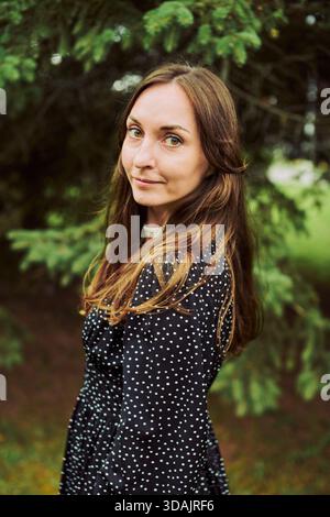 Young woman with straight brown hair and hazel eyes poses calmly in black dress with white polka dots. Background features lush greenery under soft na Stock Photo