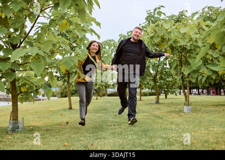 Happy young couple enjoys lively moment, holding hands and jumping while walking through vineyard with lush green grapevines on bright day. Scene capt Stock Photo