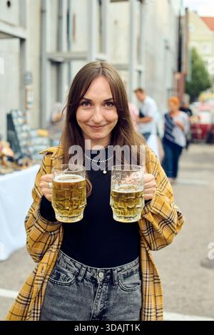 Young woman with brown hair smiles while holding two mugs of beer. She wears yellow plaid shirt over black top and stands in outdoor urban environment Stock Photo