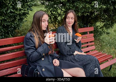 Two young women sit on red wooden bench outdoors under green bushes, holding takeaway coffee cups and engaging in lively gossip on overcast day Stock Photo
