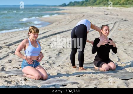 Two young women engage in pranayama breathing and body tapping exercises on sandy Baltic Sea shore under bright daylight. Calm water and green backgro Stock Photo