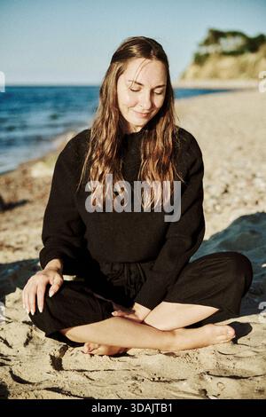 Adult woman with long brown hair and light skin sits cross-legged on sandy Baltic Sea beach. Calm expression with closed eyes, warm natural light, cli Stock Photo