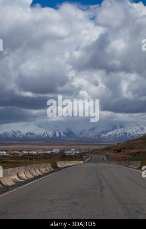 Straight paved road stretching towards a range of snow capped mountains under dark dramatic storm clouds Stock Photo