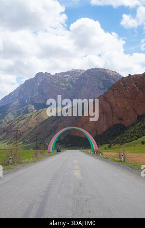 Paved road passing through a colorful rainbow-shaped archway with rugged mountains and green hills in the background Stock Photo
