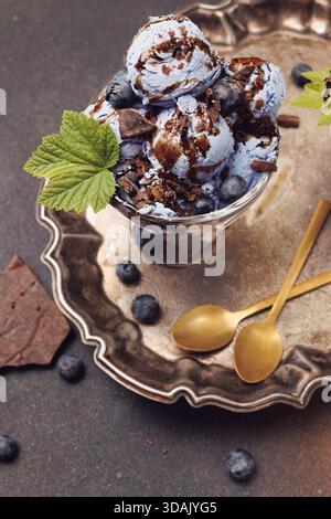 Blueberry ice cream scoops with chocolate shavings and fresh berries served in a glass dessert bowl on a rustic metal tray. Gourmet frozen dessert Stock Photo