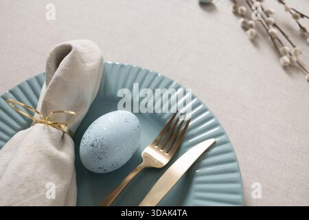 Elegant Easter table setting with blue speckled egg, gold cutlery and linen napkin on pastel plate and linen tablecloth. Soft spring decor and holiday atmosphere. Stock Photo