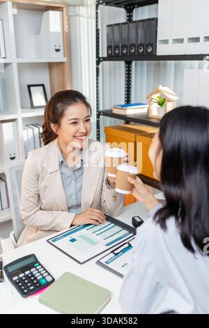 Two female colleagues take a short coffee break during their work meeting, smiling and clinking their coffee cups in a friendly gesture, showing teamw Stock Photo