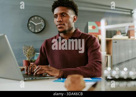 Young Adult Black Man Concentrating While Typing on Laptop in Office Workspace Stock Photo