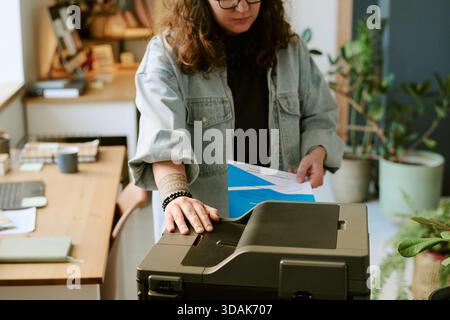 Young Adult Caucasian Woman Using Printer While Holding Documents in Office Stock Photo