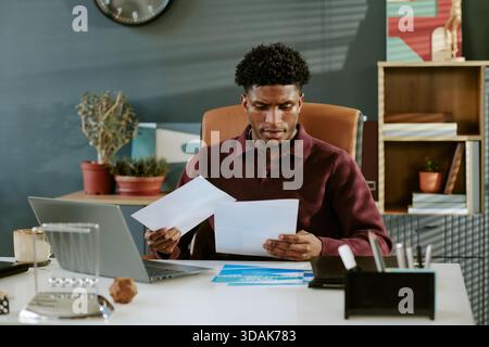 Young Adult Black Man Reviewing Financial Documents While Sitting at Desk Stock Photo