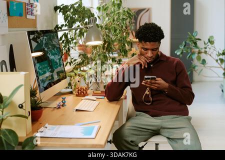 Young Adult Black Man Using Smartphone While Sitting at Modern Office Desk Stock Photo