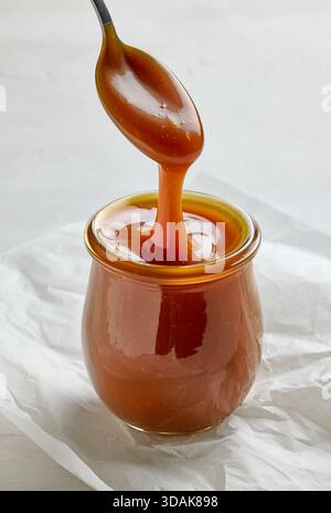freshly cooked homemade caramel sauce pouring in jar from spoon on white kitchen table background Stock Photo