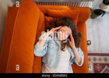 Young woman lying on an orange sofa, struggling with headache and discomfort, closing her eyes and pressing temples with her hands, indicating pain, stress, or illness Stock Photo