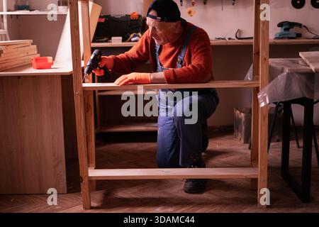 Profession, art and hobby concept. Elderly man carpenter assembling wooden shelf using electric bit drill working on wood in garage. Authentic worksho Stock Photo