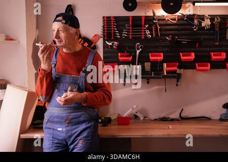 Senior Elderly Mechanic sending voice note or message using smartphone mic in workshop. copy space Stock Photo