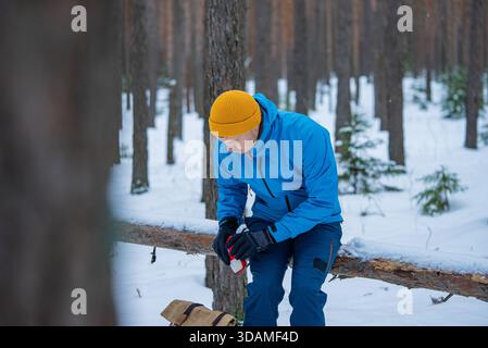 Hiker in blue jacket and yellow beanie opening a red first aid kit, sitting on a log in a snow-covered pine forest, ensuring safety during an outdoor adventure Stock Photo