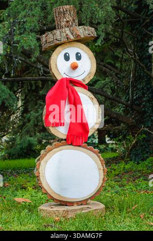 A wooden snowman stands in a garden setting. It has a wide smile and wears a red scarf. The snowman is made from tree slices and positioned on a grass Stock Photo