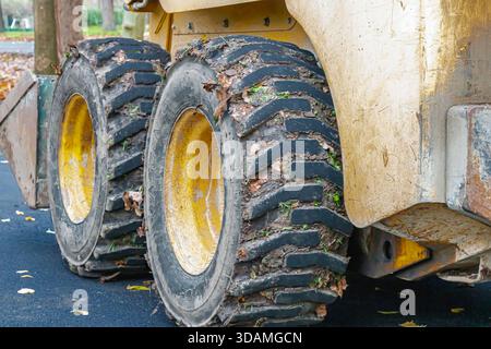 Detailed view of two large muddy tires on a yellow construction vehicle parked on a dark asphalt road. The image highlights the rugged tread pattern, Stock Photo