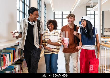 Multiethnic group of young students meeting at the library - group of diverse college students smiling, talking, and carrying books while walking in a Stock Photo
