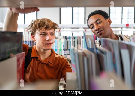 Two diverse male students are attentively browsing shelves filled with books in a brightly lit modern library, actively seeking knowledge and educatio Stock Photo