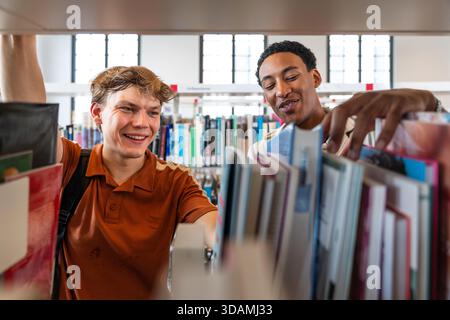 Two young diverse male students standing in a library, smiling and searching for books on shelves, representing education, knowledge, diversity, and a Stock Photo