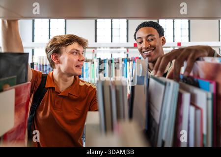Two diverse university students smiling as they browse books on a library shelf, collaborating and sharing knowledge while studying together in bright Stock Photo