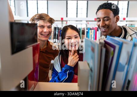 Multiethnic group of young students meeting at the library - multiethnic friends together, happily looking at books on a shelf in a modern college lib Stock Photo