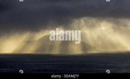 View of sunbeams piercing through the dark, stormy clouds over the sea, illuminating a distant island in a dramatic display of light and shadow, Sorva Stock Photo