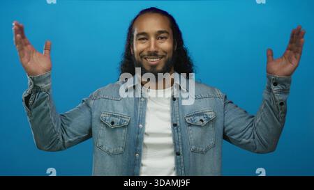 Man with hands raised palms up in blue studio wearing denim jacket and white tee and smiling face; playful joy. Stock Photo