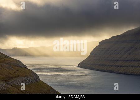 View of sun rays pierce through dramatic clouds, illuminating the tranquil waters nestled between rugged, textured cliffs, Sorvagur, Vagar, Faroe Isla Stock Photo