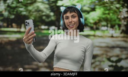 Young hispanic woman with cat ear headphones holds smartphone and beckons hand in forest; friendly invitation. Stock Photo