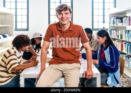 Group of young diverse students actively engaged in collaborative learning and studying at a table in a bright library, while one male student is smil Stock Photo