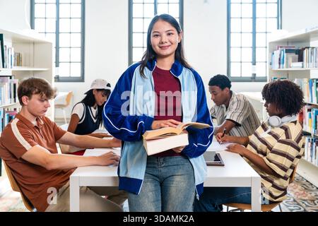 Diverse university students studying together in a modern library, multi-ethnic group smiling and focused as they hold textbooks and collaborate at a Stock Photo