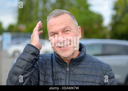 Cheerful portrait of a mature man in a black jacket smiling warmly and touching his hair outdoors on a bright day. Stock Photo