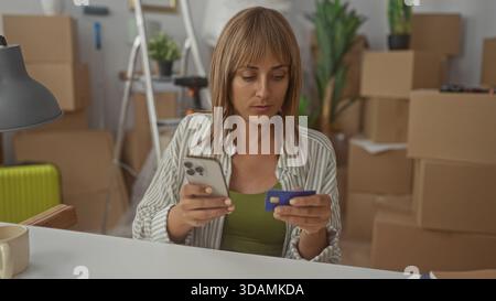 Woman holding smartphone and creditcard in building among packed boxes and green suitcase checking payment on phone; moving planning focus. Stock Photo