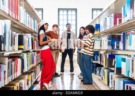 Multiethnic group of young students meeting at the library - diverse young student friends discussing learning topics at university library aisle, sha Stock Photo