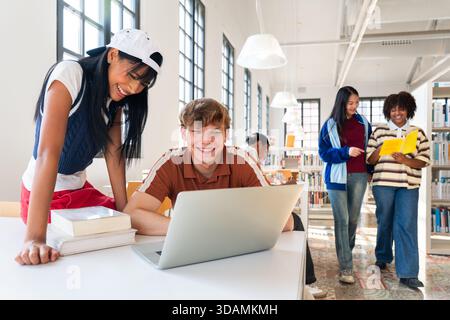 Multiethnic group of young students meeting at the library - diverse students working together in a university library, using a laptop, reading books, Stock Photo