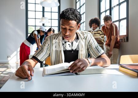 Group of high school students collaborating and focused on learning in a bright school library, with one teen deeply engrossed in reading an open book Stock Photo