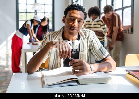 Young male student holding a pen and looking at the camera while studying from an open book at a desk in a university library with other students in t Stock Photo