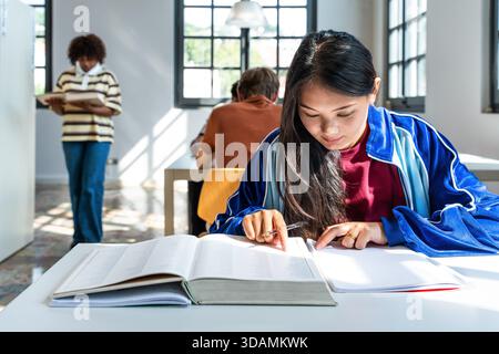 Young woman concentrating on a large open book at a table, highlighting concepts of education, learning, research, and academic pursuit within a moder Stock Photo