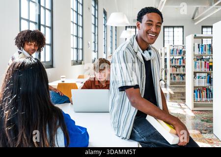 Students collaborating and studying together in a modern library, with a smiling young man in the foreground holding books and looking at the camera, Stock Photo