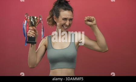 Woman holds trophy with fist raised in studio against a vivid red backdrop, smiling broadly; pride celebration. Stock Photo