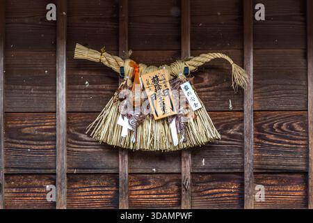 An Ise-Shimekazari hangs on a wooden wall in Oharai-machi, Ise, Mie Prefecture, Japan. The protective amulet, woven from rice straw, carries two wooden plaques: on the left a blessing from Ise Jingū, on the right the characters for "safety and well-being of the household", plus dried plants, a gourd as a good-luck symbol, and strips of shide paper. The Ise-Shimekazari is a regional variant of the shimenawa purification symbol found throughout Japan and is hung across the city as a New Year's and protective charm. Stock Photo