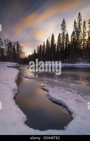 View of icy river reflecting the pastel sunset between snow-covered banks and silhouetted trees, a serene winter scene, Kuusamo, North Ostrobothnia, F Stock Photo