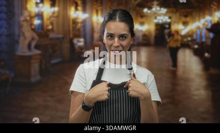 Young brunette woman wearing a striped apron points finger to object while standing in ornate museum building lobby; welcoming hospitality. Stock Photo