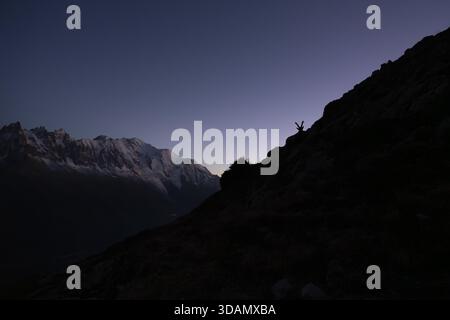 View of a small figure standing on a dark slope with hands up, silhouetted against the backdrop of snowy mountains and a gradient sky, Chamonix-Mont-B Stock Photo