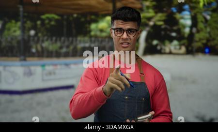 Young hispanic man in glasses taking notes in a park setting, wearing a red shirt and apron, captured outdoors with a thoughtful expression, enhancing Stock Photo