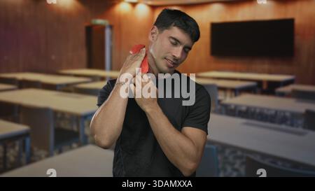 Man holds red hotpack to neck while wincing from muscle ache in classroom with empty desks and chairs; relief. Stock Photo